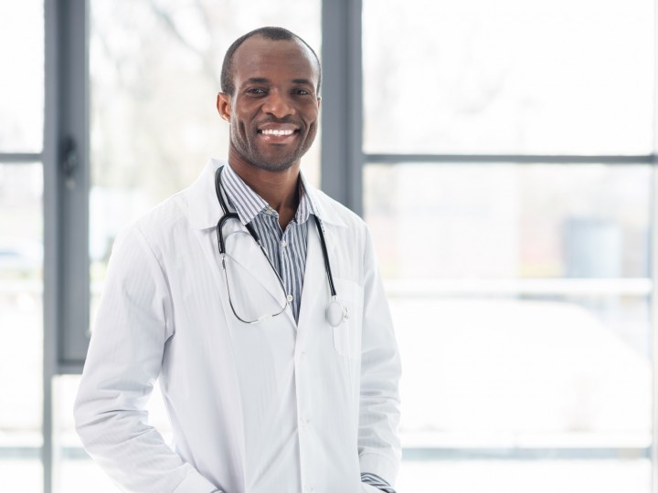 A doctor smiles and stands in front of window with white coat and stethoscope on