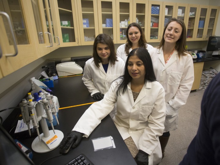 smiling students standing behind Dr.Malik in the lab while using a pipette